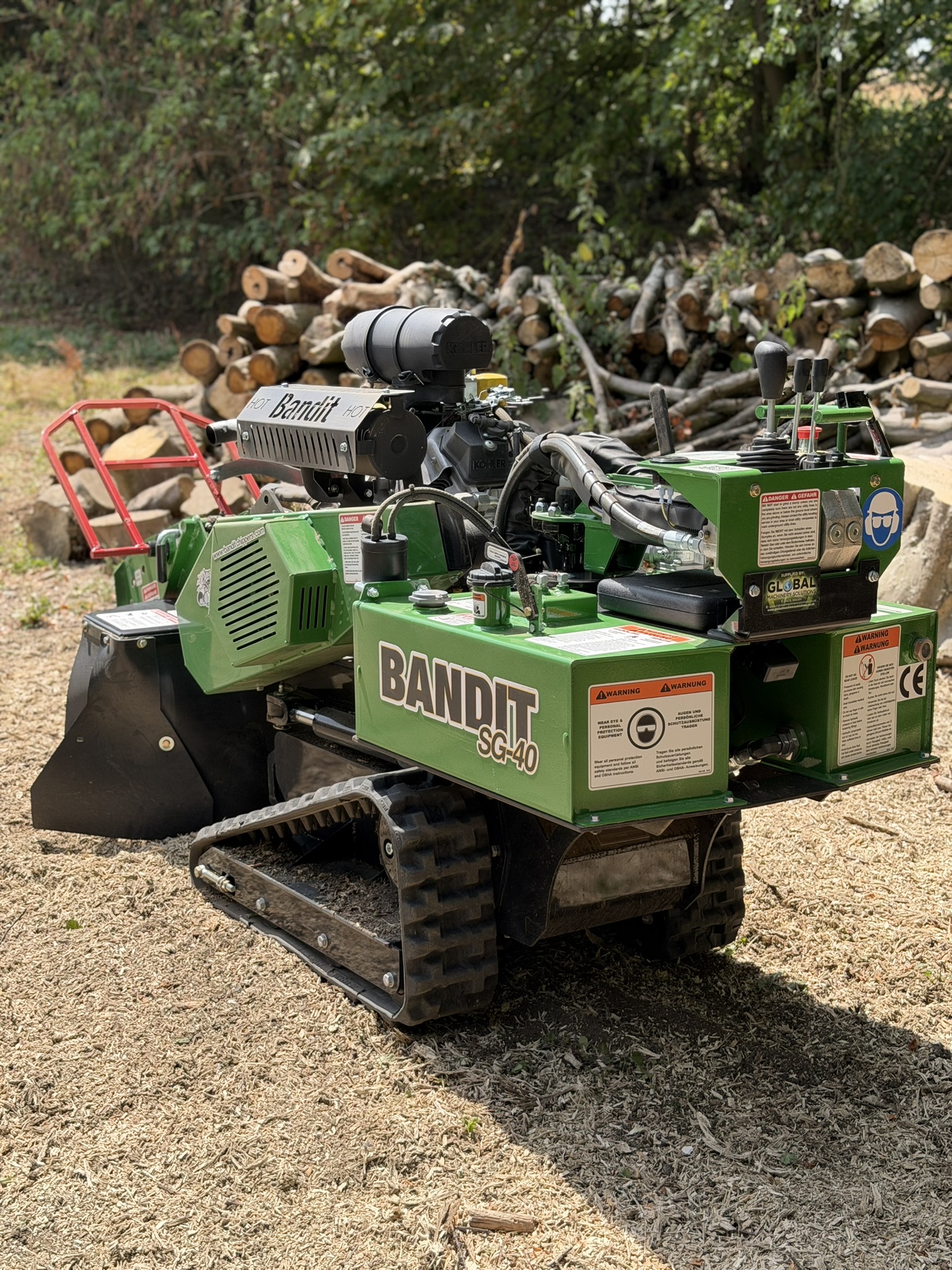 The green Bandit SG-40 tracked stump grinder parked on a bed of wood chippings with a large log pile behind on the Much Hadham site