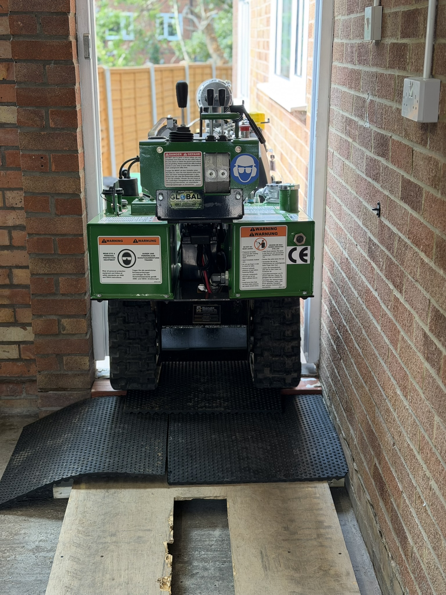 A green Bandit SG-40 tracked stump grinder driven head-on through a narrow doorway between brick walls, running over rubber matting and plywood boards laid to protect the ground into the Loughton garden