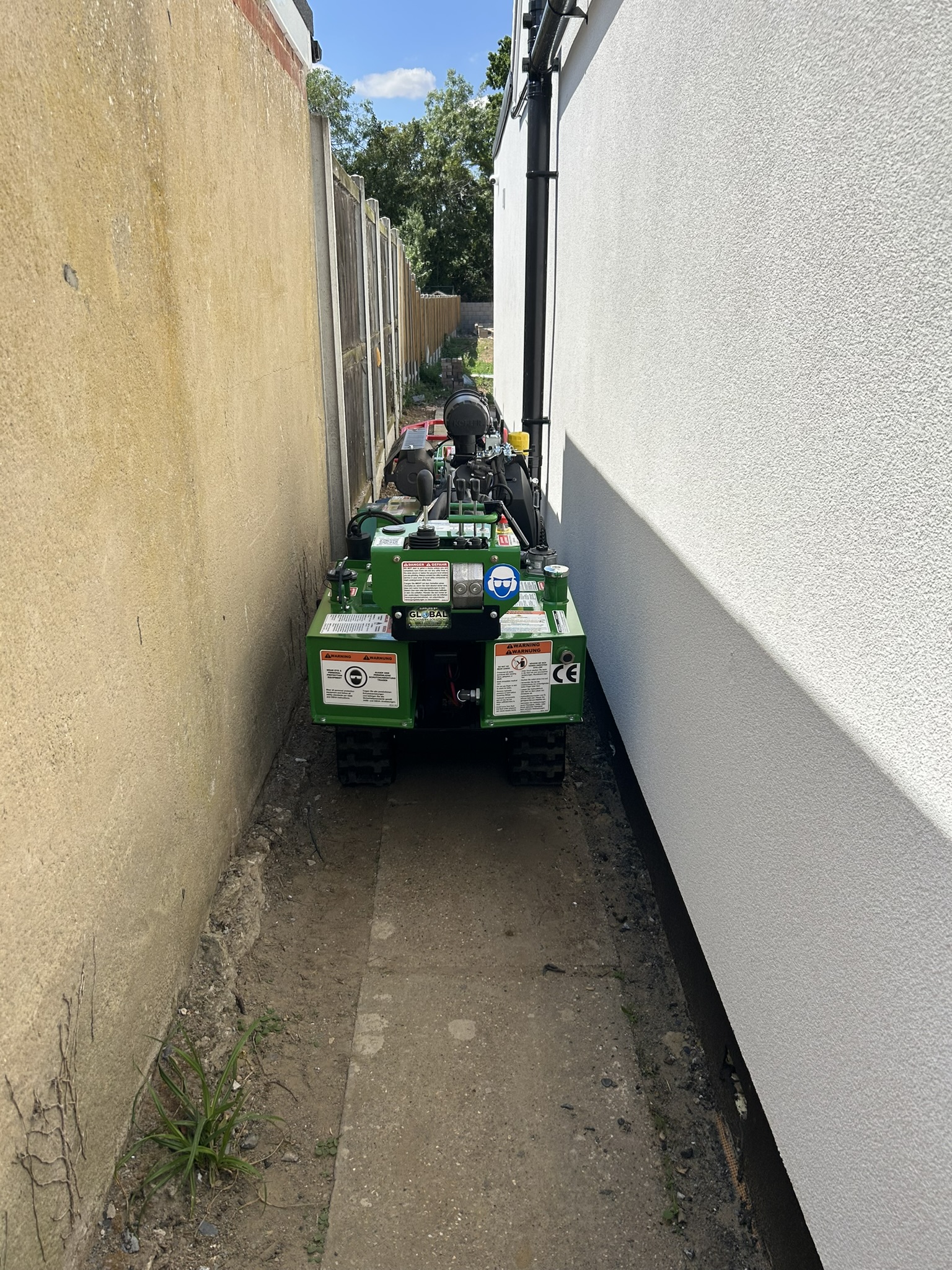 A green tracked Bandit stump grinder driven down a very narrow concrete alleyway between a rendered house wall and a boundary wall, with only a few inches of clearance on either side
